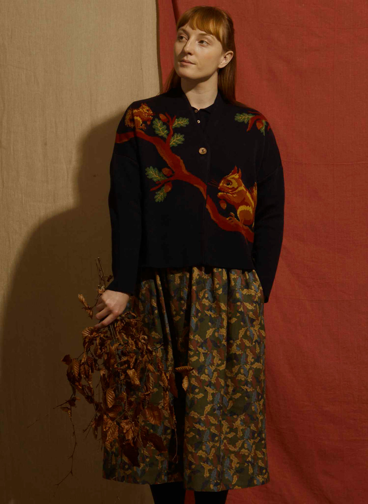 A woman stands against a split beige and rust-red fabric backdrop, wearing a dark navy cardigan with a red fox and tree design on the chest, paired with a patterned skirt. She holds a bundle of dried leaves.