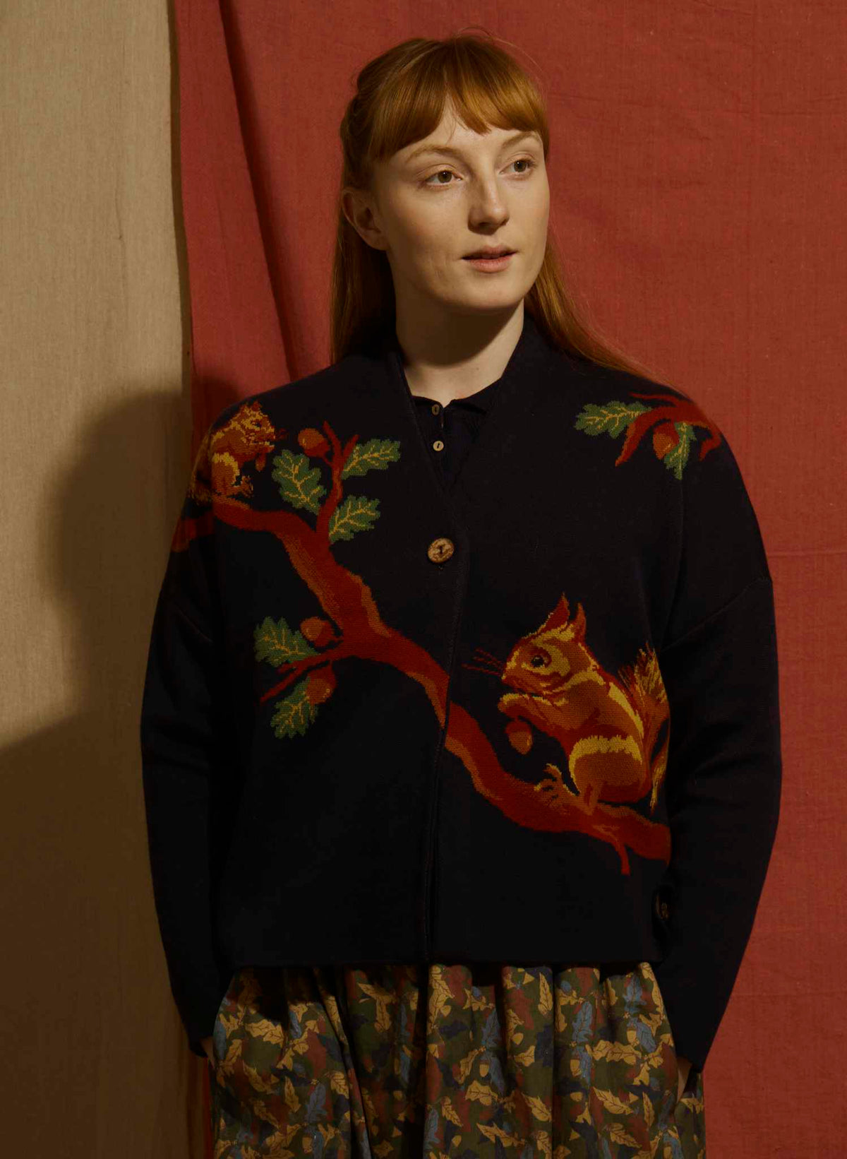 A woman with red hair stands against a red fabric backdrop wearing a dark navy cardigan with a large embroidered squirrel and tree design on the front, paired with a patterned skirt.