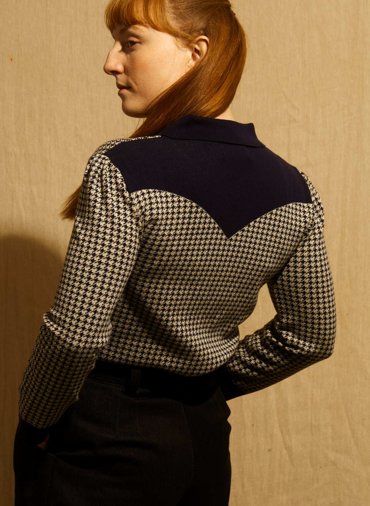 A women wearing a navy and beige houndstooth jacket with dark navy blue trim on the collar, cuffs, hem, and button details, and navy trousers, shown against a wooden background. The back profile shows a vintage cowboy-style motif.