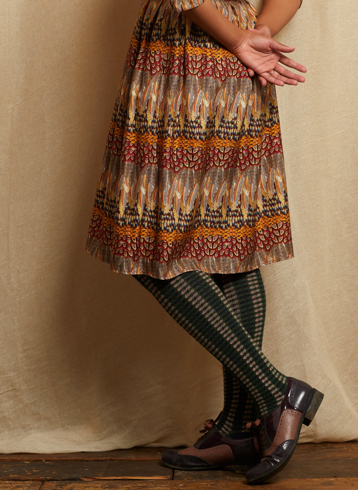 A person wearing a knee-length feather patterned dress with horizontal bands of rust red, mustard yellow, beige, and dark brown, paired with dark green patterned tights and dark brown leather shoes, standing against a draped beige fabric backdrop.