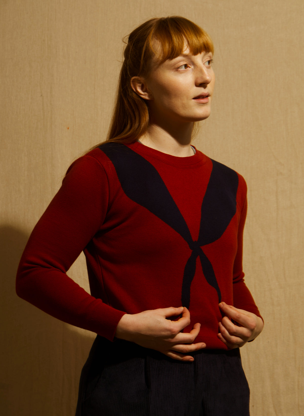 A woman wearing a cropped crewneck sweater in deep red with a large dark navy blue scarf motif across the chest - and dark navy trousers - against a wooden backdrop.