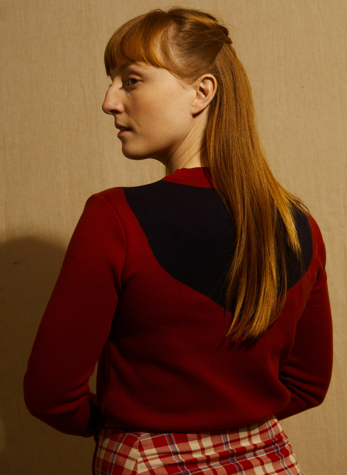 A woman wearing a cropped crewneck sweater in deep red with a large dark navy blue scarf motif across the chest - and a red/white check kilt skirt - againsts a wooden backdrop.