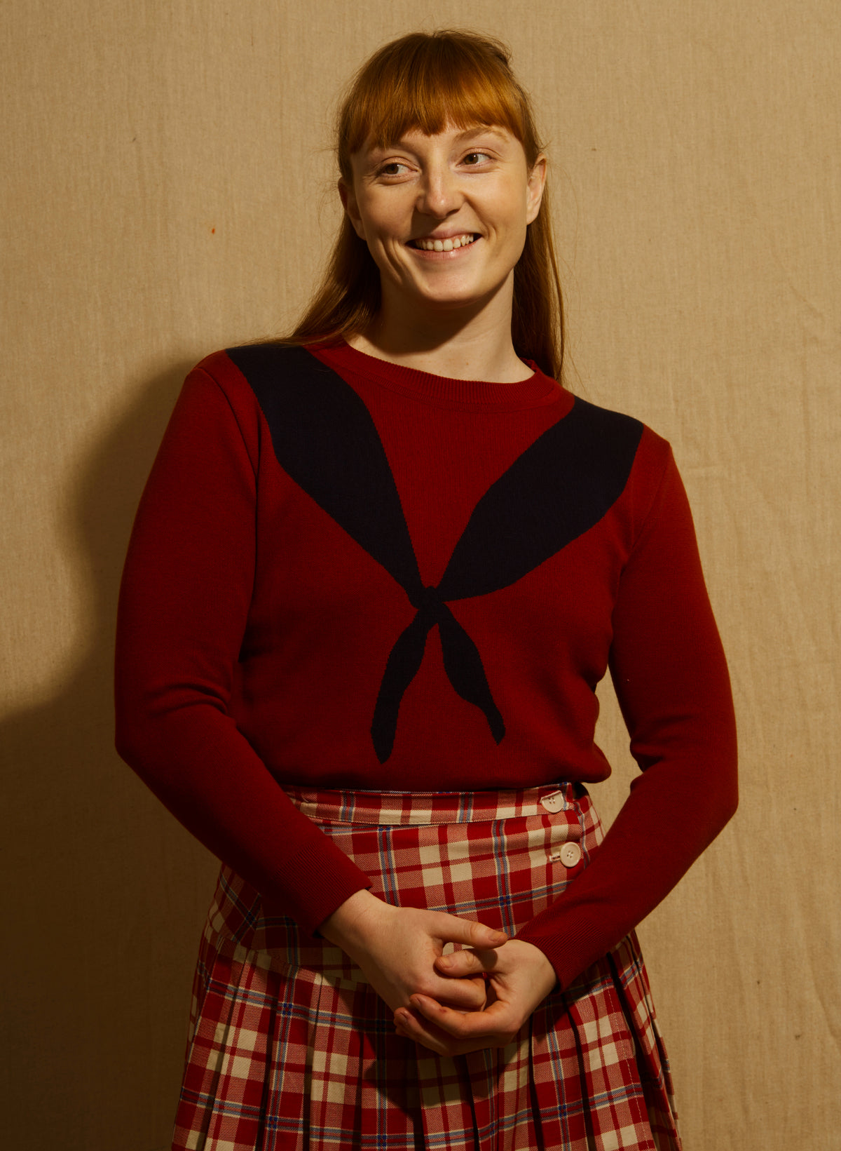 A woman wearing a cropped crewneck sweater in deep red with a large dark navy blue scarf motif across the chest - and a red/white check kilt skirt - againsts a wooden backdrop.