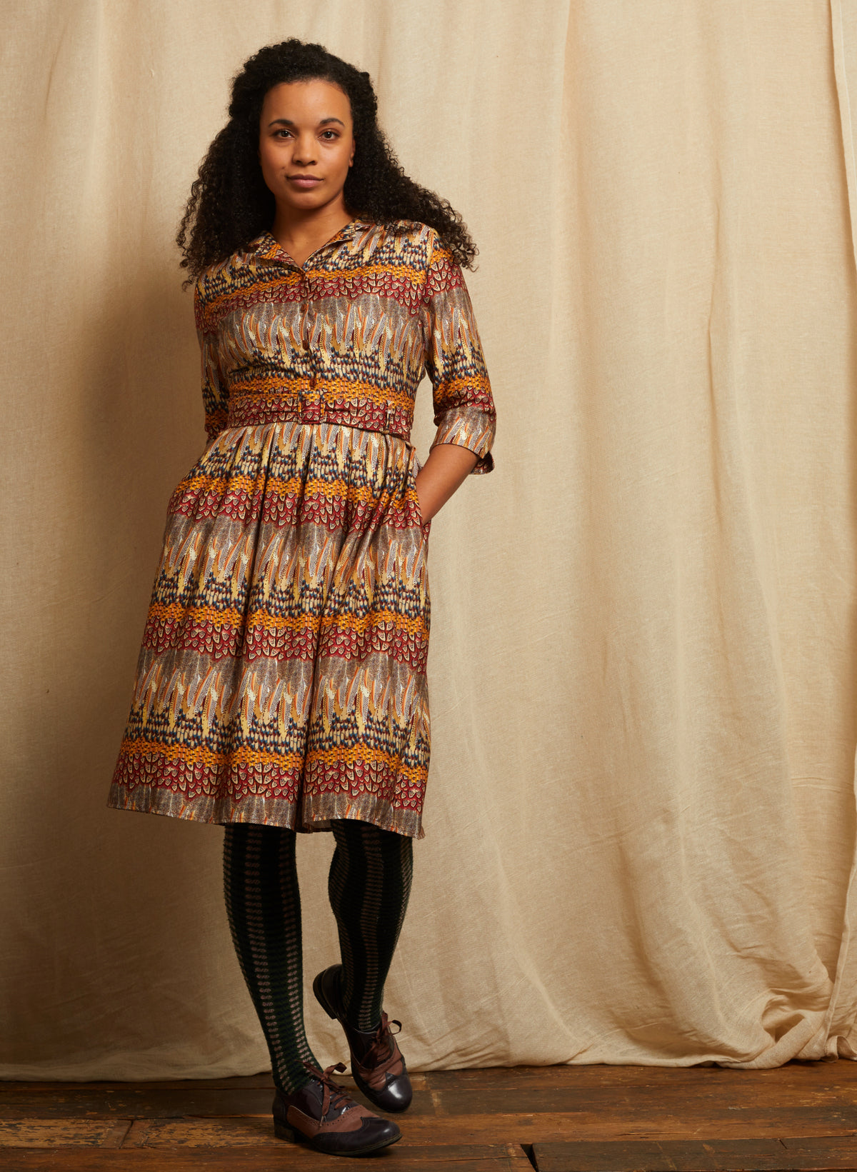A woman stands in front of a draped beige fabric backdrop wearing a patterned midi dress with three-quarter sleeves, a collared neckline, and a gathered waist. The dress has an intricate multicolor feather print featuring red, orange, yellow, blue, and black motifs on a cream base.