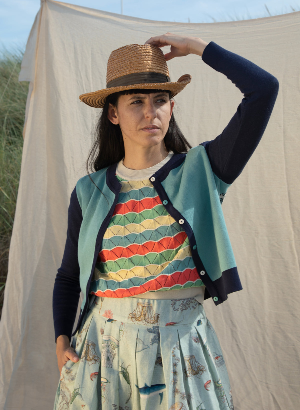 A picture of a woman from the front, she is wearing a cardigan featuring a mineral blue body with navy arms. Underneath is a multicoloured short sleeve knitted top. The woman is wearing a summer hat and she is looking out to sea. 
